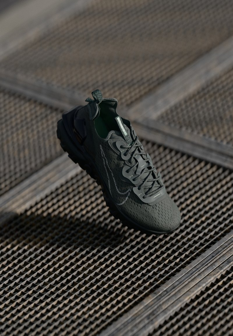 Zielony but sportowy o teksturowanej powierzchni, skomplikowanym wzorze sznurowadeł i wyraźnej gumowej podeszwie, zawieszony nad wzorzystą metalową siatką.
