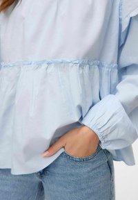 Blusa azzurra chiaro a maniche lunghe con dettagli di rouches arricciati in vita e sui polsini, abbinata a jeans in denim azzurro chiaro.