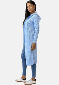 Długi, jasnoniebieski sweter na drutach z wzorem warkocza, kapturem i prążkowanymi krawędziami; noszony na obcisłych dżinsach i trampkach.