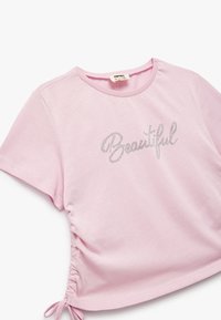Camiseta de manga corta de color rosa claro con lados fruncidos y letras de strass plateado que dicen "Hermosa". Material de algodón suave.