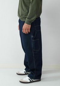 Pantalons cargo en denim bleu marine avec poches latérales et coutures jaunes contrastantes, associés à des sneakers blanches et grises.