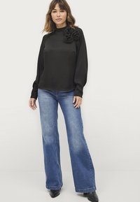 Zwarte langemouwen top met een hoge kraag en drie stoffen rozen op de schouder, gecombineerd met hooggetailleerde blauwe wijde jeans.