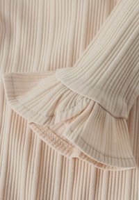 Top de manga larga color durazno claro con textura acanalada vertical y puños con volantes. Tejido suave con un leve brillo y detalles fruncidos en las mangas.