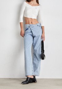 Vaqueros de denim de corte holgado en azul claro con cintura media y costuras en contraste. Top corto blanco ajustado con frente fruncido y mangas largas. Mules negras.
