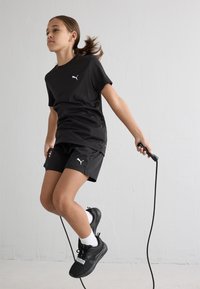 Melna sporta apģērba komplekts sastāv no īsām piedurknēm krekla un šortiem, abiem rotātiem ar baltu logo. Subjekts dejo ar virvi, ģērbies melnās kurpēs.