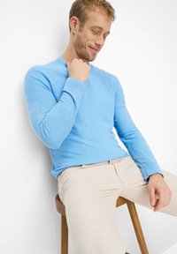 Lyseblå trøje med lange ærmer i en struktureret strik, kombineret med beige bukser, siddende på en træskammel mod en hvid baggrund.