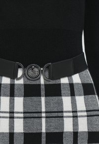 Zwarte riem met zilveren ronde gesp van schakels over een zwart-witte geruite rok en een zwarte geribbelde trui.