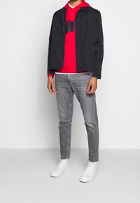 Czarna lekka kurtka na czerwonej bluzie z kapturem z granatowym wzorem, zestawiona z szarymi dżinsami i białymi tenisówkami. Prosty i nowoczesny design.