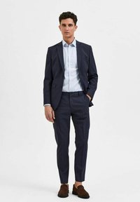 Marinefarget skreddersydd dress med slim-fit bukser, hvit og blåstripete skjorte under, og brune loafers. Glatt stoff, klare linjer og moderne design.