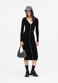 Vestido preto canelado com um decote em V profundo e fecho éclair frontal, combinado com um boné cinza e uma mala macia cinza, usado com meias pretas e sapatos.