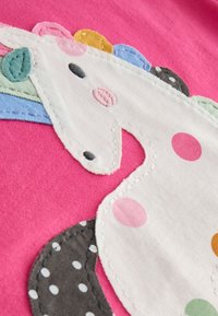 Motif de licorne brodé sur tissu rose ; présente une crinière multicolore, des pois et diverses textures. Fond blanc avec des détails de couture.