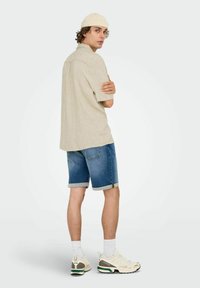 Lysbeige kortærmet skjorte, denim shorts med opkrøllede manchetter, hvide sneakers med grønne detaljer, og en lys kasket, set fra bagfra.