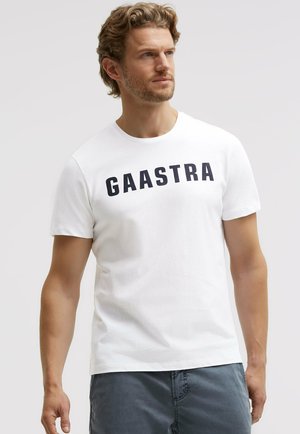 Mann mit lockigem Haar, der ein weißes T-Shirt mit dem Schriftzug "GAASTRA" und blaue Hosen trägt, schaut seitlich vor einem schlichten Hintergrund.