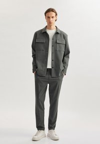 Camisa gris de botones con dos bolsillos en el pecho, combinada con pantalones grises a juego y zapatillas blancas. Textura suave, corte relajado.