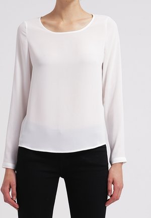 Vrouw draagt een effen witte blouse met lange mouwen en een ronde hals, gecombineerd met zwarte broek, staand tegen een neutrale achtergrond.