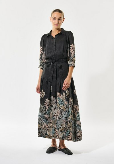 Robe noire à manches longues avec broderies florales dans des couleurs pastel. Présente un devant à boutons, une taille cintrée et une longueur au genou. Chaussures plates noires.
