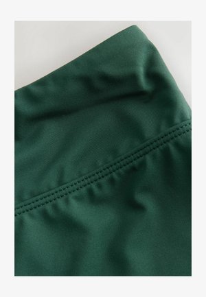 Mørkegrønt stof med en glat overflade. Har et bredt taljebånd og flade sømme. Materialet virker strækbart og fugttransporterende.