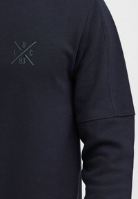 Mørk marineblå hettegenser med teksturert overflate, som har en ton-i-ton logo-design på brystet og en avslappet passform med en litt senket skuldersøm.