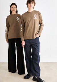 Brown long-sleeve shirts with white text on sleeves, paired with black wide-leg pants and blue striped jeans, worn with black shoes.