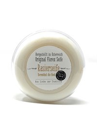 White, circular shaving soap in a clear, plastic container. Label features black and gold text, detailing origin and product name. Smooth texture.