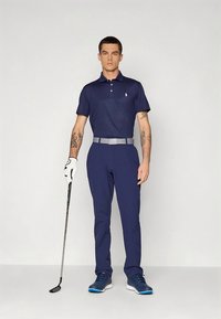 Tmavě modré polo tričko s krátkými rukávy, tmavě modré kalhoty se štíhlým střihem, šedý pásek, bílá golfová rukavice a modré sportovní boty. Minimalistický design s logem.