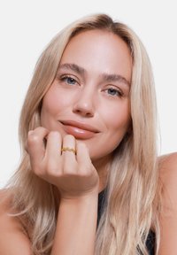 Goldring mit wellenförmigem Design und kleinen Perlen, am Zeigefinger getragen. Die Hand ruht auf dem Kinn vor einem weißen Hintergrund.