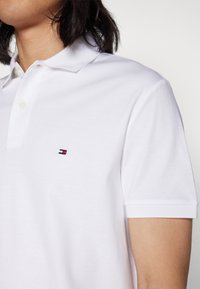 Maglietta polo bianca con una texture morbida, dotata di una placca con tre bottoni e un piccolo logo ricamato in rosso, bianco e blu sul petto.