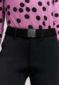 Ceinture noire avec boucle en plastique attachée autour d’un pantalon noir, portée avec une chemise rose à gros pois noirs.
