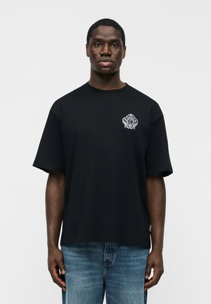 Mladý muž s tmavou pokožkou, oblečený v čiernom oversize tričku s bielym emblémom na hrudi a modrých rifliach, stojaci čelom dopredu.