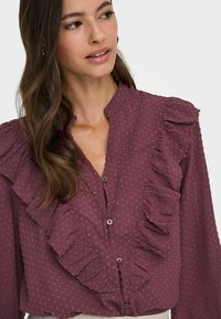 Blouse bordeaux avec des pois texturés, présentant un col haut, un devant froncé et une fermeture à boutons. Manches longues avec des accents froncés.