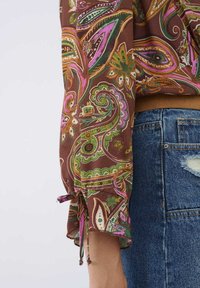 Bruine blouse met een paisleydessin in groen, roze en geel. Beschikt over geraffineerde mouwen met strikken en een textuurstof.