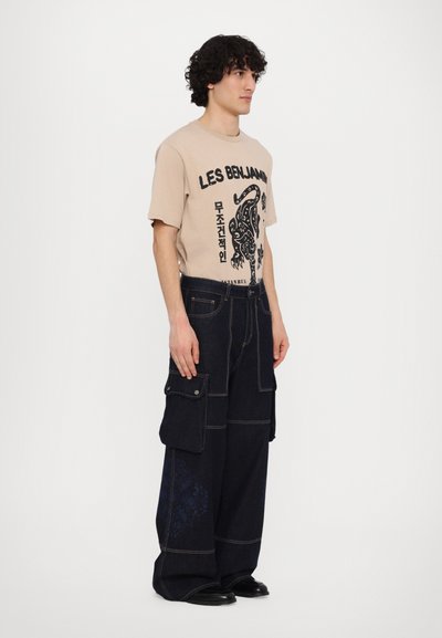 T-shirt beige avec impression graphique et pantalon cargo large bleu foncé à poches multiples avec des accents floraux bleus.