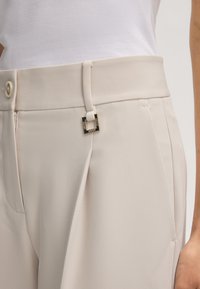 Primo piano di pantaloni sartoriali color crema con pieghe, passanti per cintura, bottone, cerniera e targhetta quadrata in metallo del marchio vicino alla vita.