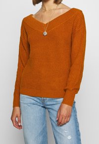 Gestrickter oranger Pullover mit weitem V-Ausschnitt und langen, leicht aufgeblasenen Ärmeln. Mit gerippten Bündchen und Saum, kombiniert mit ausgefransten blauen Jeans.