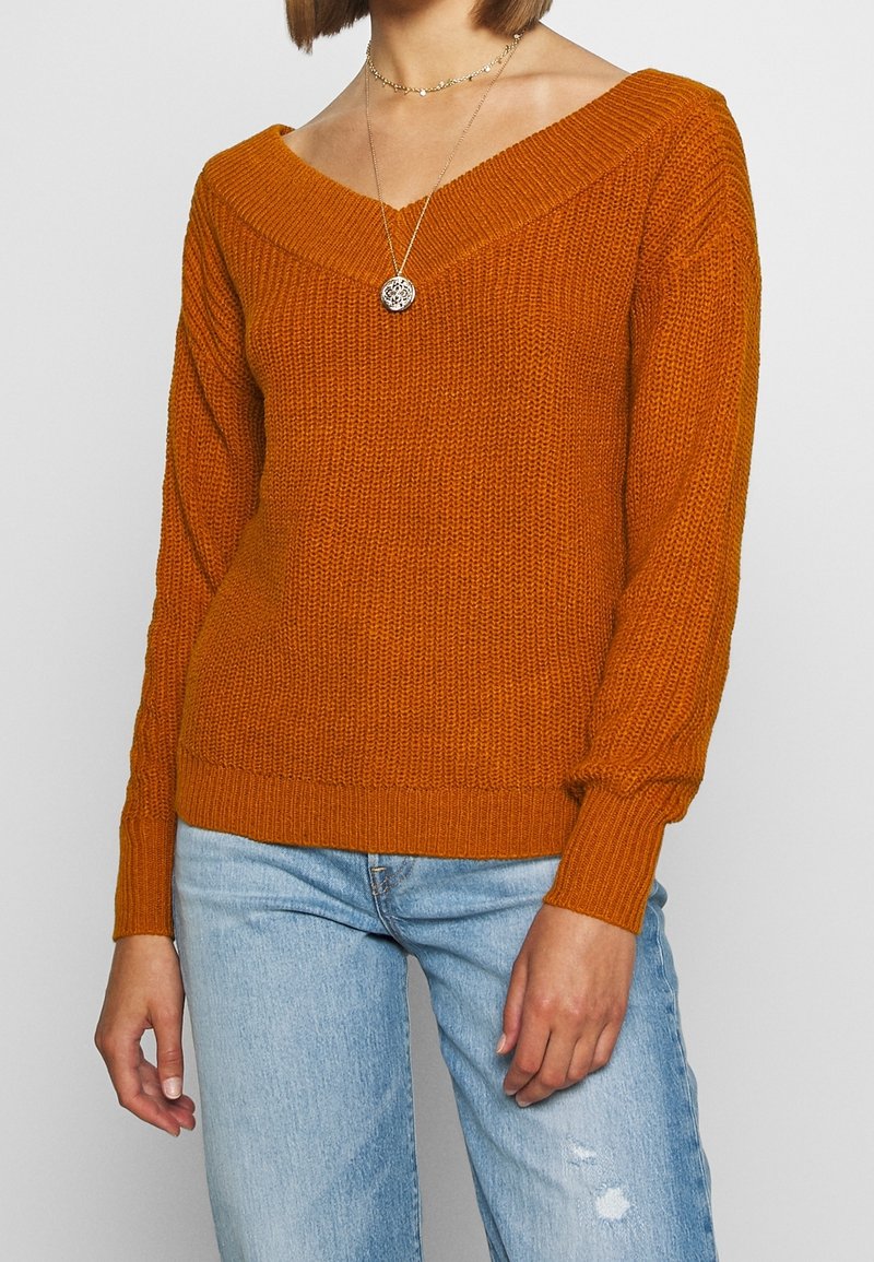 Gestrickter oranger Pullover mit weitem V-Ausschnitt und langen, leicht aufgeblasenen Ärmeln. Mit gerippten Bündchen und Saum, kombiniert mit ausgefransten blauen Jeans.