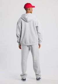 Sudadera gris con capucha amplia, puños acanalados y pantalones de chándal grises a juego. Gorra roja llevada al revés. Diseño casual, tejido suave, ajuste relajado.