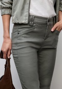 Grå åtsittande byxor i slätt tyg med dragkedja, bältesöglor och sidofickor. En brun mockaväska hålls i höger hand.