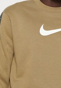 Khakifärgad sweatshirt med rund halsringning, med en stor vit Nike-logotyp på bröstet och ett texturerat tyg som visar lätt stretch.