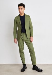 Abito verde oliva con giacca su misura e pantaloni tapered, abbinato a una maglietta a girocollo blu navy e scarpe stringate nere su uno sfondo neutro.