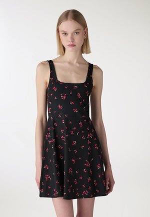 Giovane donna che indossa un abito nero senza maniche con stampe di ciliegie rosse, in piedi contro uno sfondo chiaro uniforme.