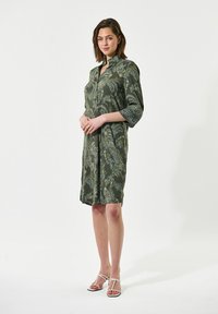 Grünes gemustertes Hemdkleid mit Dreiviertelärmeln, Kragen und lockerer Passform, kombiniert mit weißen Riemchensandalen.