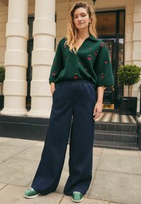 Pulover verde tricotat cu aplicații în formă de cire�șe, asortat cu pantaloni largi bleumarin și adidași verzi. Poziționat în fața unui fundal arhitectural clasic.