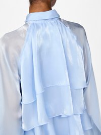 Hellbluse mit Kragen. Verfügt über geschichtete, ruffled Details und eine schimmernde Stofftextur.