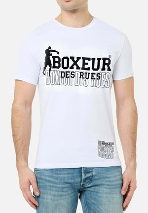 Uomo che indossa una maglietta bianca con silhouette nera di boxe e testo "BOXEUR DES RUES" e "THE FIGHTER BRAND" sopra jeans azzurri.