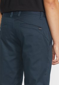 Navy blue pants with a slim fit, featuring two back pockets, a small brand tag, and a smooth, textured fabric.
