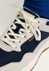 Baskets avec un dessus beige et marine, en matériaux en suède et en tissu, lacets crème et logo orange. Présente une semelle intermédiaire texturée blanche et une semelle en caoutchouc.