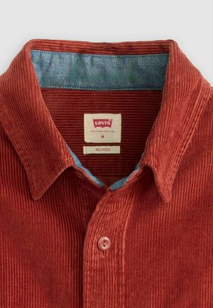 Camisa de pana color óxido con forro de cuello azul, corte holgado, con dos botones y una etiqueta roja con la marca. Tejido con textura acanalada.