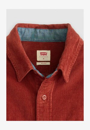 Roestkleurig corduroy overhemd met een blauwe kraagvoering, ontspannen pasvorm, voorzien van twee knopen en een rood label met merkteken. Getextureerde ribstof.