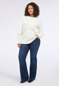Maglione in maglia di colore crema con dettagli in frange lungo la vita, abbinato a jeans a zampa di elefante blu scuro e scarpe con tacco a blocco marroni.