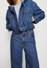 Oříznutá džínová bunda ve středně modré barvě s šedou kapucí, vybavená dvěma předními kapsami a stahovací šňůrkou. K tomu široké džíny v podobném modrém odstínu.